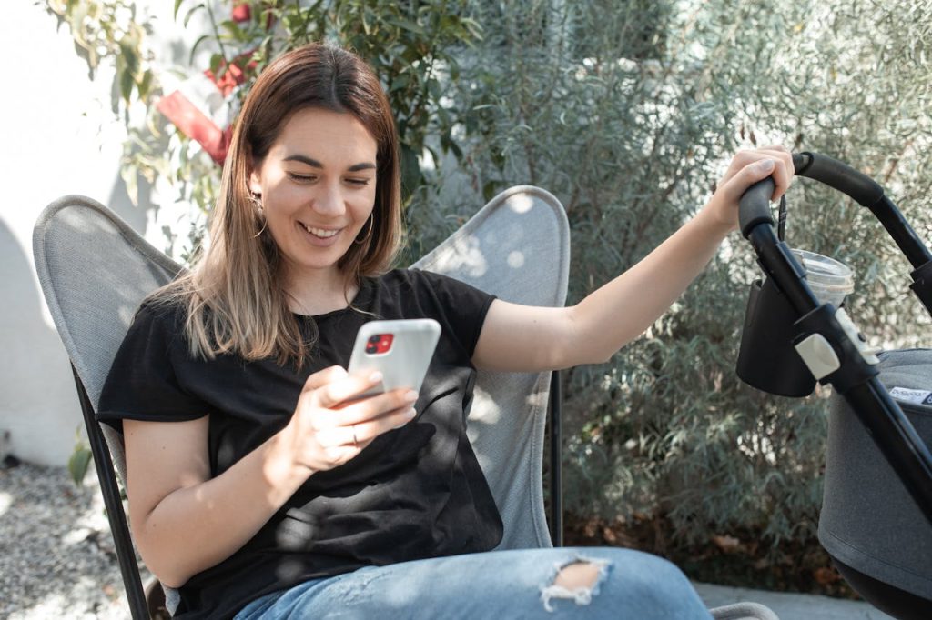 pexels-photo-6849496 Woman seated outdoors, smiling while using smartphone beside a stroller.